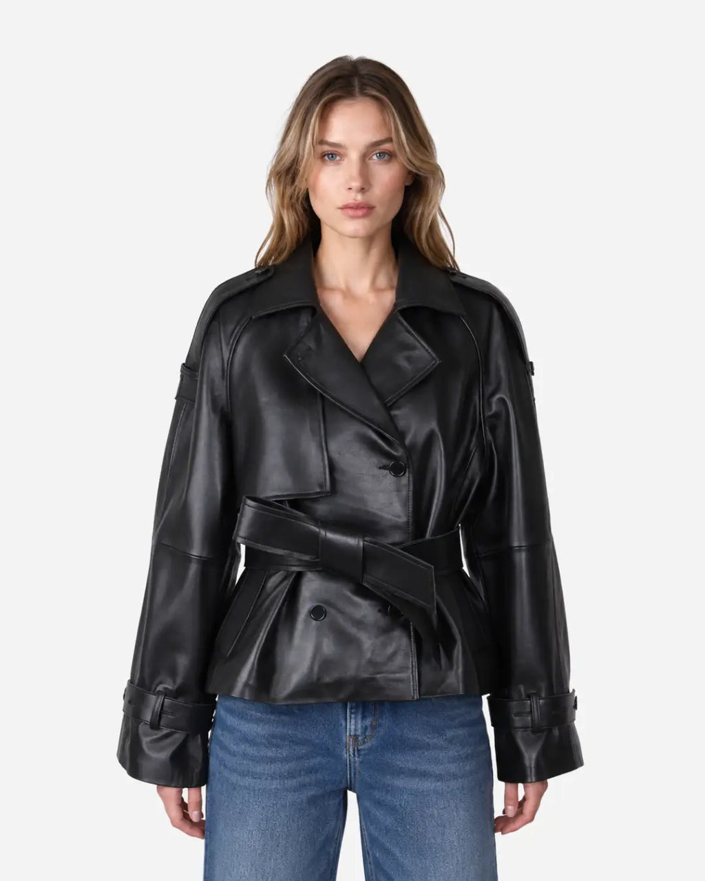 A woman modeling a black leather belted jacket with wide lapels and cuffed sleeves, paired with blue jeans, against a light gray studio background.