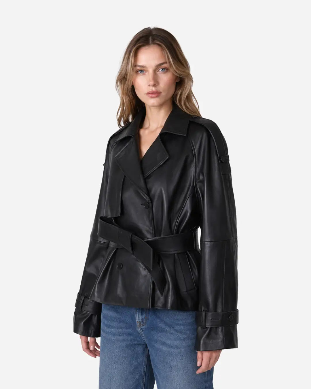A woman modeling a black leather belted jacket with a collar and wide lapels, paired with blue jeans, against a light gray studio background.