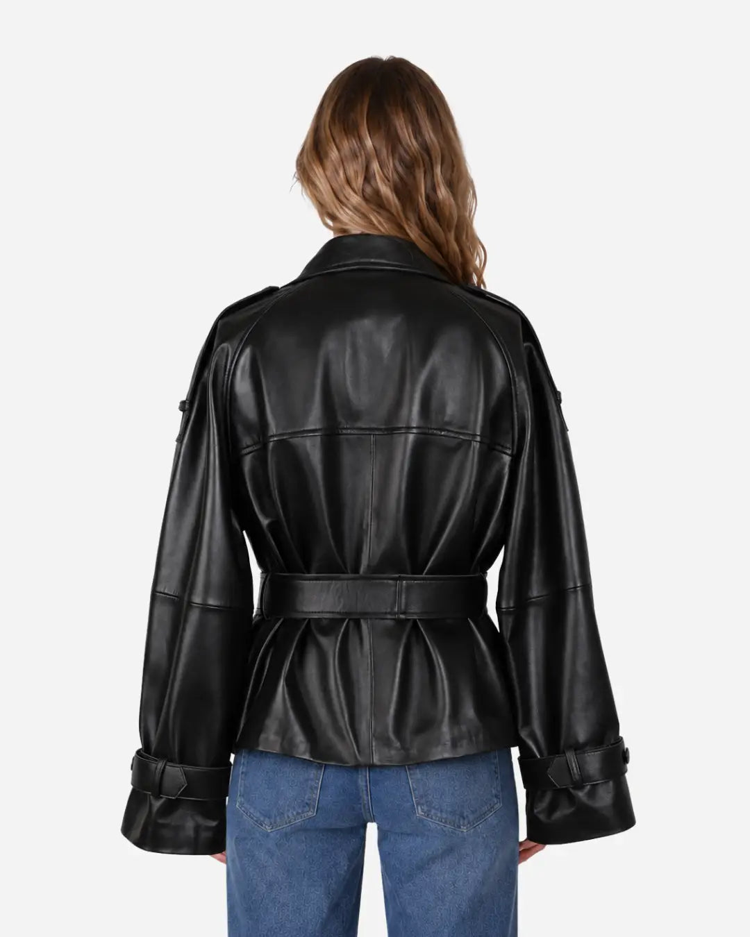 A woman viewed from the back wearing a black faux-leather jacket with a belted waist and cuffed sleeves, paired with blue jeans, against a plain light gray background.