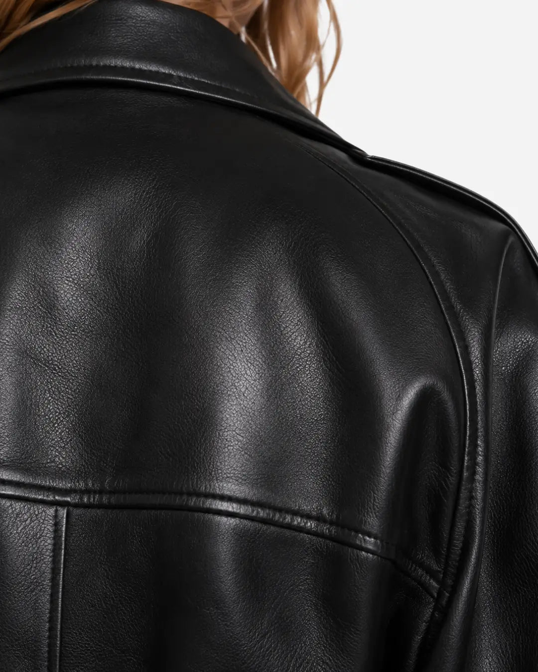 Close-up of a black leather jacket on a model, showing the back collar, shoulder, and textured leather surface against a light gray background.